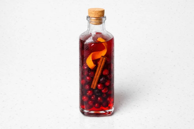 Bottle of deep red Christmas infused vodka with cranberries, cinnamon stick, and orange peel visible through the glass