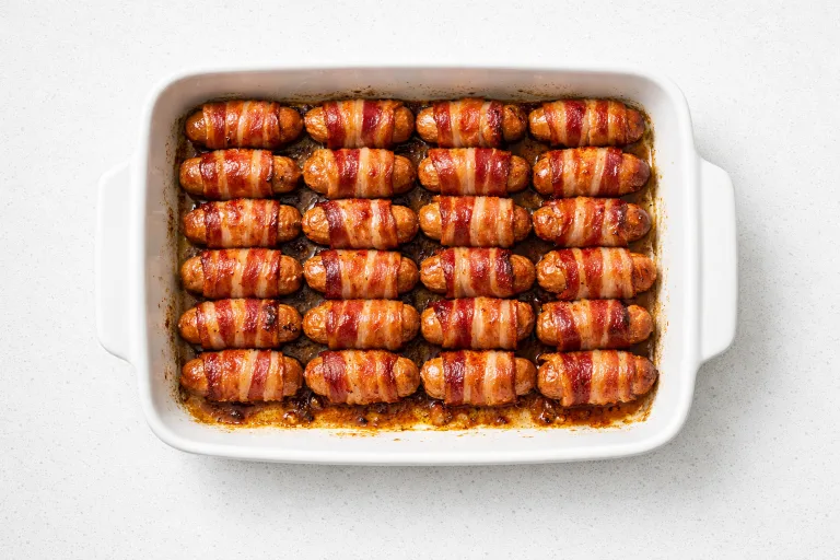 Golden crispy pigs in blankets in a white roasting dish, chipolatas wrapped in streaky bacon, caramelised and glistening