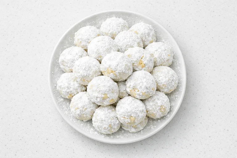 Mexican wedding cookies (polvorones) piled on a white plate, dusted with powdered sugar to resemble snowballs