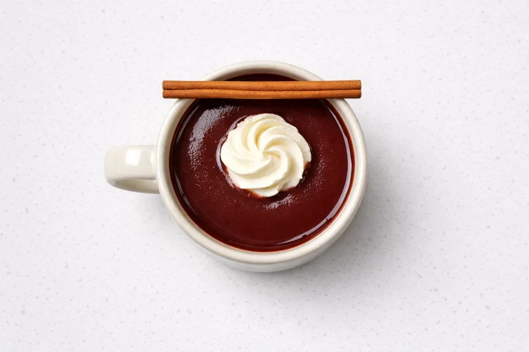 Overhead view of a ceramic mug of red wine hot chocolate with whipped cream and a cinnamon stick on white countertop