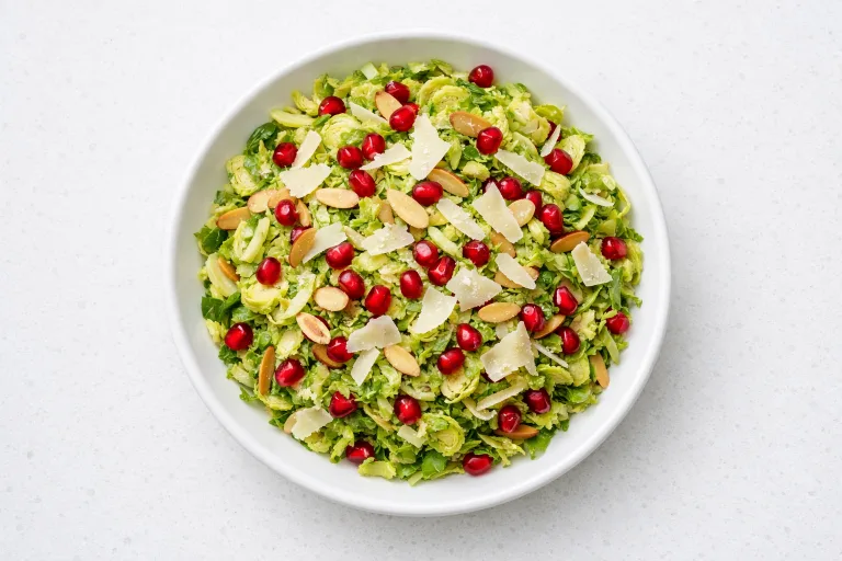 Shaved brussels sprout salad with pomegranate seeds, toasted almonds, and Parmesan in a white bowl