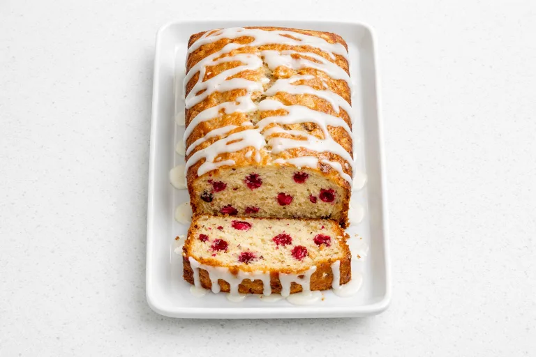 Sliced cranberry orange bread with orange glaze on a white ceramic plate, showing bright red cranberry pieces throughout the golden crumb