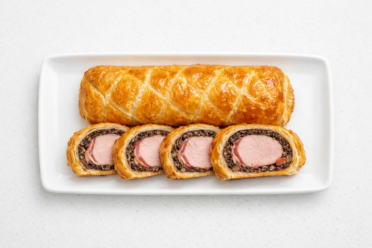 Sliced pork wellington with mushroom duxelles on a white platter showing golden pastry and pink tenderloin center