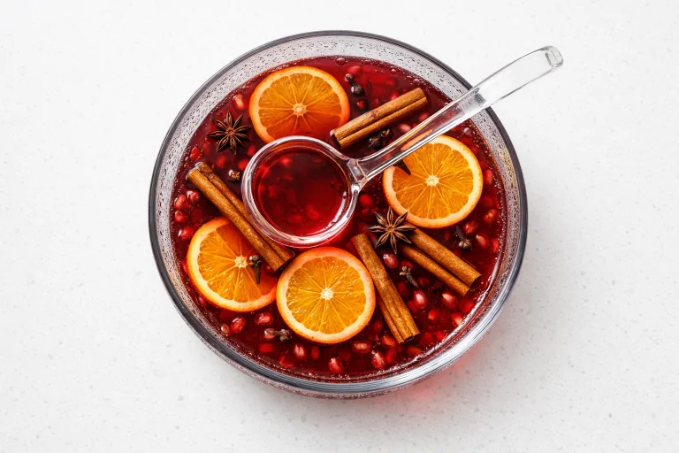 Spiced pomegranate punch in a clear glass punch bowl with orange slices and cinnamon sticks floating on the surface