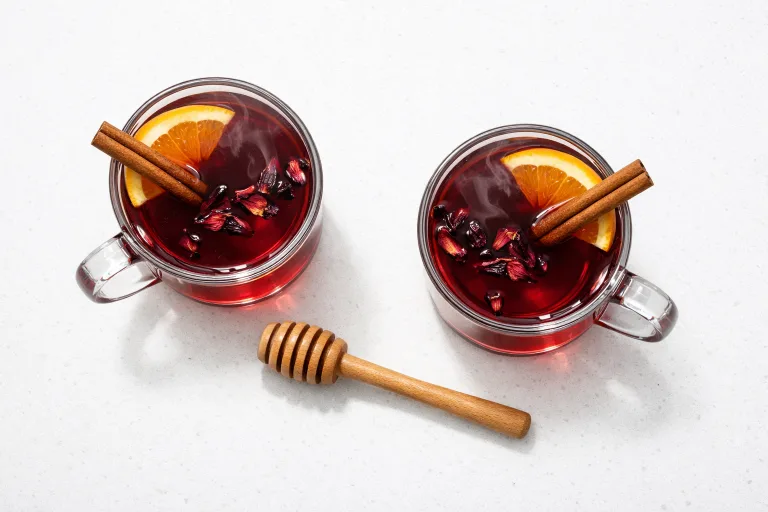 Two glass mugs of deep red hibiscus hot toddy with cinnamon sticks and orange slices on the rim, steaming on a white surface