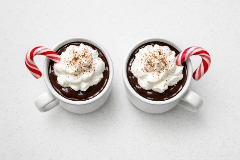 Two mugs of homemade peppermint hot chocolate topped with whipped cream and candy canes, overhead view